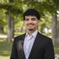 Samuel is standing in front of some trees. He has short black hair and is wearing a black blazer.