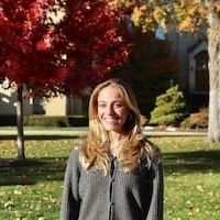 Dena is standing on campus. She has long blonde hair and is wearing a grey jacket.